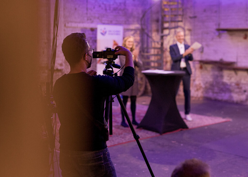 Das Bild zeigt den Frankfurter Fotografen und Unternehmer Fritz Philipp, der eine Kamera bedient. Er befindet sich am Set des Zonta Benefits Livestreams im Schaerferaum, dem Mietstudio für Bad Homburg, Fankfurt und Umgebung. Im Hintergrund sieht man eine der Zonta Damen zusammen mit einem Moderator von den Clown Doktoren