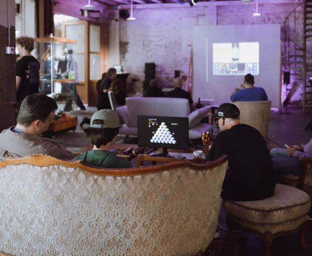 Mehrere Besucher sitzen auf Sofas und Sesseln und spielen an historischen Computersystemen. Im Hintergrund werden Inhalte auf eine Leinwand projiziert. Die Aufnahme entstand während der Ausstellung des Homecomputer Museum Frankfurt im Studio Schärferaum in Bad Homburg.